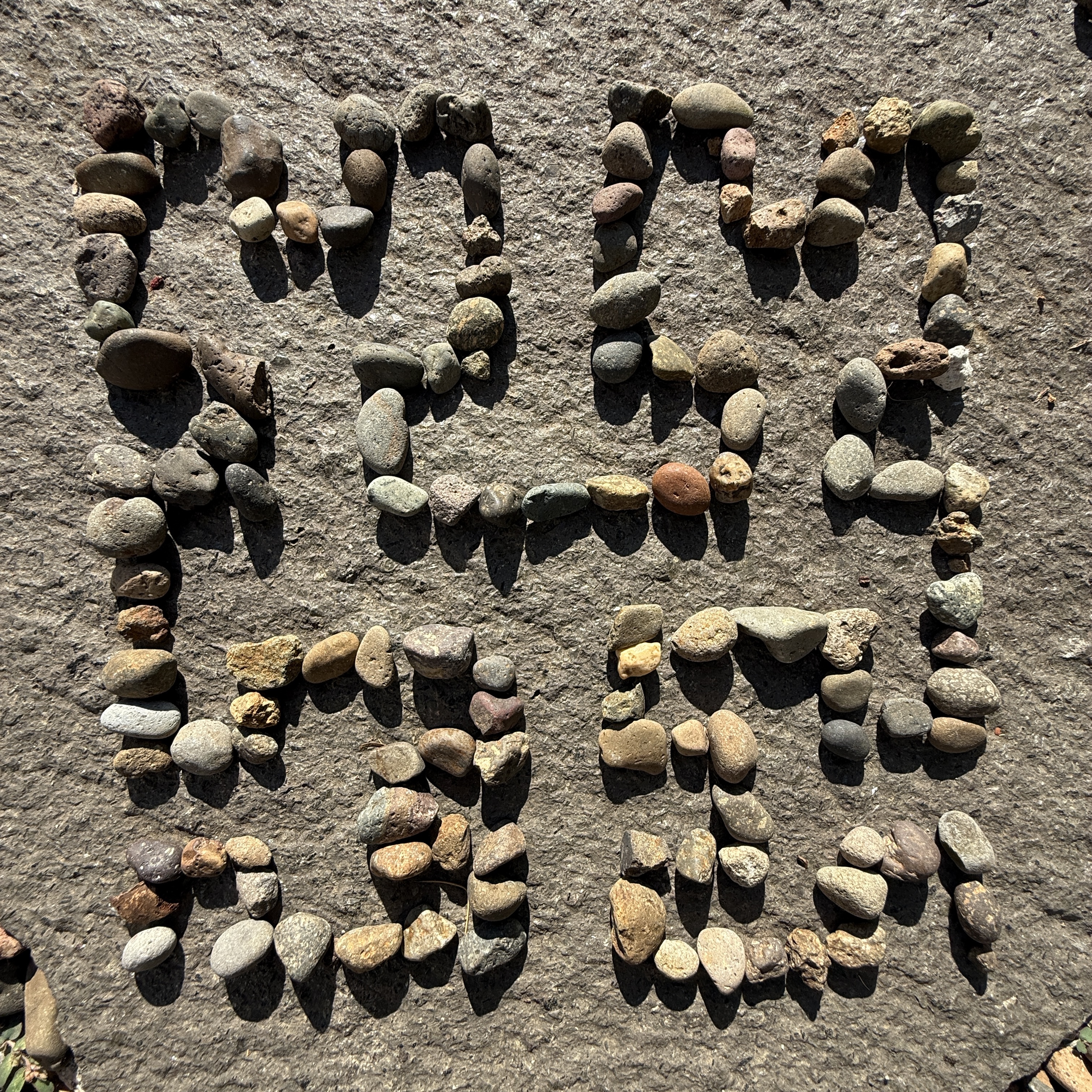 A Hilbert curve drawn with pebbles on a flat rock surface. The pebbles have many shades and shapes and the Hilbert curve looks like 4 squarish cat-heads with ears where the top two are facing up and the bottom two are facing in and they are all drawn with a single line of pebbles (like line art) so they are connected. Shadows point downward from horizontal pebbles showing the morning light.