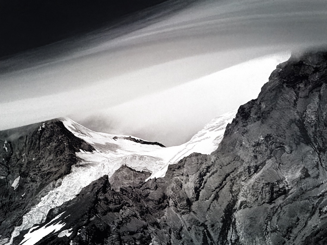 A lenticular cloud over a glacier on Mount Rainier a parabolic bowl with a lid of cloud. The image is high contrast, black and white. The lenticular cloud is smooth like silk and contrast to a rough and ragged structure of mountain and ice.